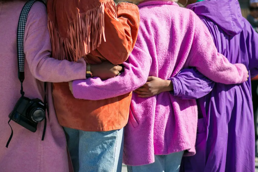Four women with their backs facing the camera. They have their arms around each other, offering support. They are wearing pink, purple and orange coats.
