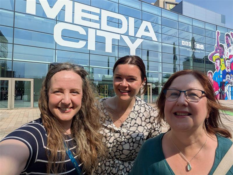 Three women stand outside the MediaCityUK studios in the sunshine